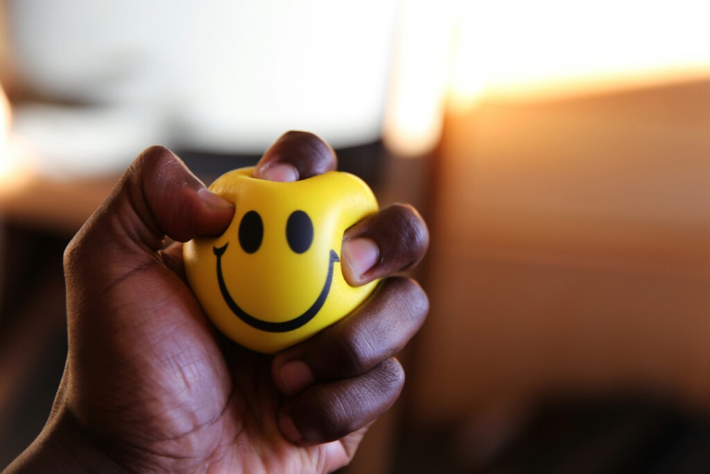 persons hand squeezing a small yellow stress ball with a smiley face on it.
