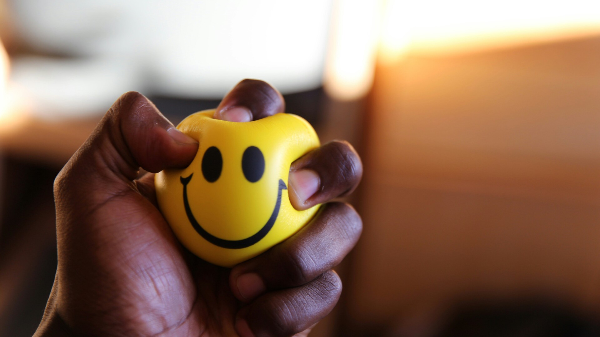 persons hand squeezing a small yellow stress ball with a smiley face on it.